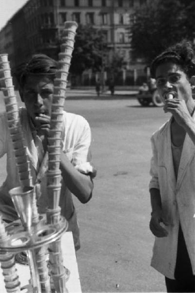 Milano. Due gelatai con il carretto - uno di essi mangia un cono - in primo piano pile di coni