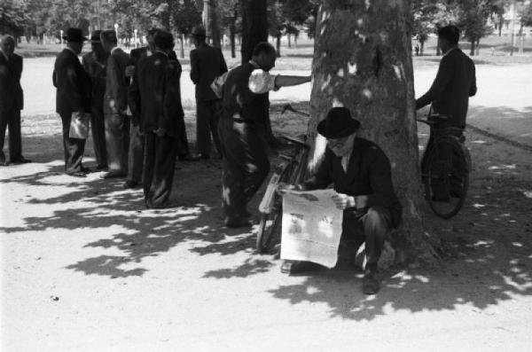 Milano. Parco Sempione. Gruppi di persone che discutono nei pressi della "Fontana dell'acqua marcia" - uno di essi siede sotto un albero a leggere il giornale