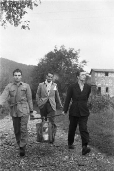 Movimento Resistenza Partigiana. Due partigiani camminano per una strada di campagna trasportando una valigia appesa ad un bastone - un altro uomo li segue