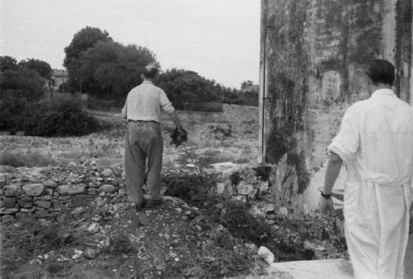 Diano Marina. Dottore con camice bianco e anziano fra il muro della cascina e i campi