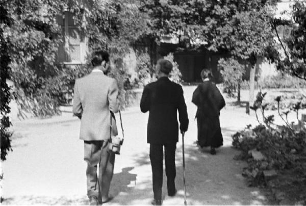 Roma. Don Luigi Sturzo ripreso di spalle mentre passeggia in un parco con una donna e un fotografo