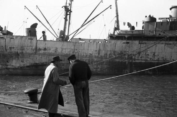 Savona. Porto. Un marinaio e un uomo in impermeabile discutono sulla banchina - sullo sfondo una nave ancorata