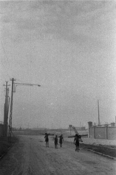 Porto Marghera. Bambini portano assi di legno sulle spalle, camminando a fianco di binari