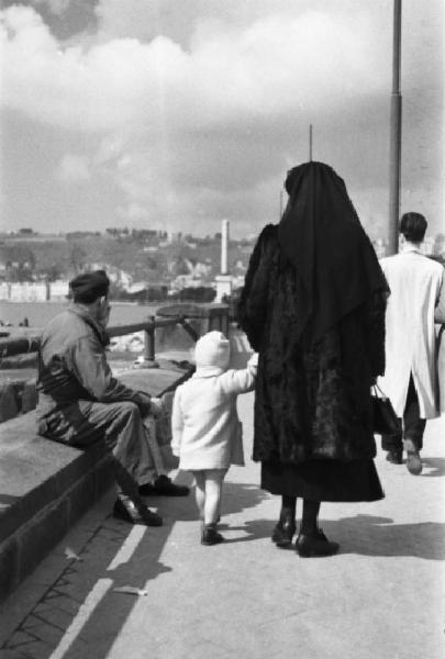 Italia Dopoguerra. Napoli. Una donna di spalle, vestita di nero e con velo, tiene per mano una bambina. Un militare seduto su un muretto fuma una sigaretta