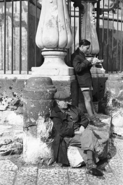 Italia Dopoguerra. Napoli. Un uomo anziano seduto e un ragazzo appoggiato a una colonna mentre consuma in piedi il pranzo