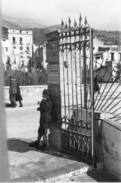 Italia Dopoguerra. Napoli. Un ragazzo appoggiato al muro d'ingresso di una cancellata guarda i passanti