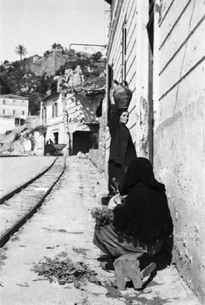 Italia Dopoguerra. Terracina. Due donne lungo la strada: una seduta pulisce delle erbe, l'altra trasporta una brocca d'acqua