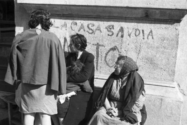 Italia Dopoguerra. Napoli. Donne sedute in prossimità di un muro inneggiante alla Casa Savoia