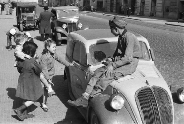 Italia Dopoguerra. Napoli. Un ragazzino seduto sul cofano di un automobile parla con delle bambine