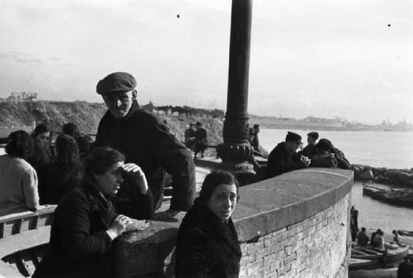 Italia Dopoguerra. Pozzuoli. Due donne e un uomo anziano accanto a un muretto presso il porto