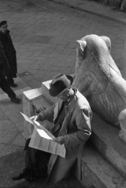 Italia Dopoguerra. Napoli. Uomo con cappello seduto accanto a una scultura in pietra legge il giornale