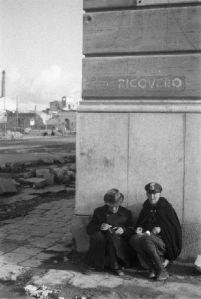 Italia Dopoguerra. Palermo. Due uomini anziani seduti accanto ad un muro recante la scritta "RICOVERO"