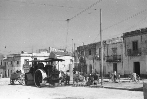 Italia Dopoguerra. Mondello. Gruppo di bambini guarda un coetaneo mentre si arrampica su un monumento. Una macchina schiacciasassi ferma lungo la strada. Abitazioni dullo sfondo