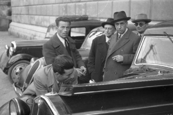 Palermo. Ritratto di gruppo - quattro uomini in abito elegante, mentre un uomo ripara un automobile