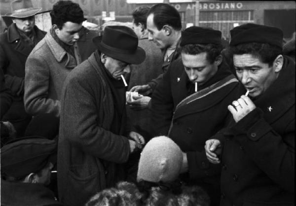 Italia Dopoguerra. Milano. Un gruppo di uomini lungo la strada si accende una sigaretta