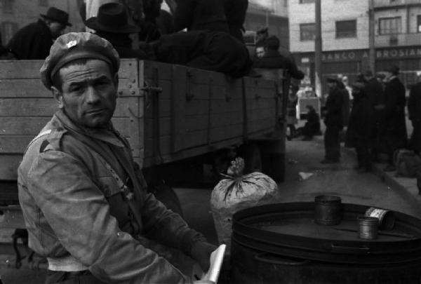 Italia Dopoguerra. Milano. Un venditore di caldarroste ritratto mentre persone si accalcano per salire su un camion