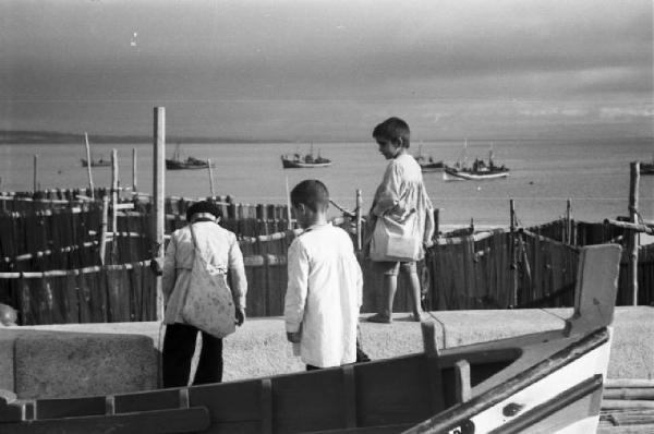 Portogallo. Cascais. Tre bambini in piedi su un muretto - al di là le reti dei pescatori e barche nell'oceano