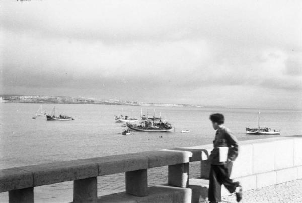 Portogallo. Cascais. Ragazzo con quaderno sotto il braccio osserva la spiaggia al di là di un muretto - barche nell'oceano