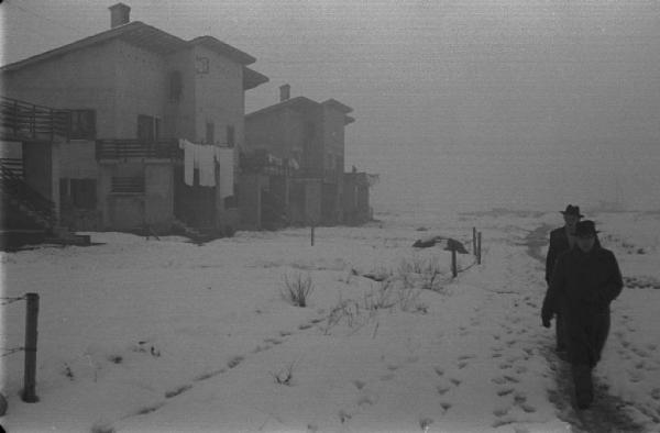 Due uomini camminano lungo un sentiero in un campo innevato a lato di abitazioni