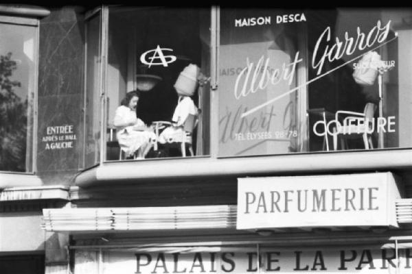 Parigi. La vetrina al primo piano di un negozio di parrucchiere. Una donna sotto il casco, un'altra esegue la manicure