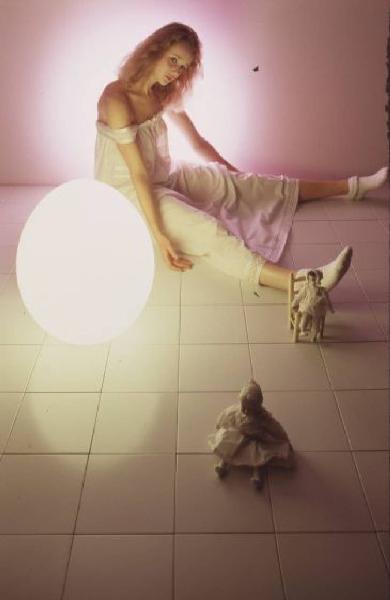 Ragazza vestita da bambola posa su un pavimento a lato di una lampada tonda - ai suoi piedi rimangono appoggiate due bambole