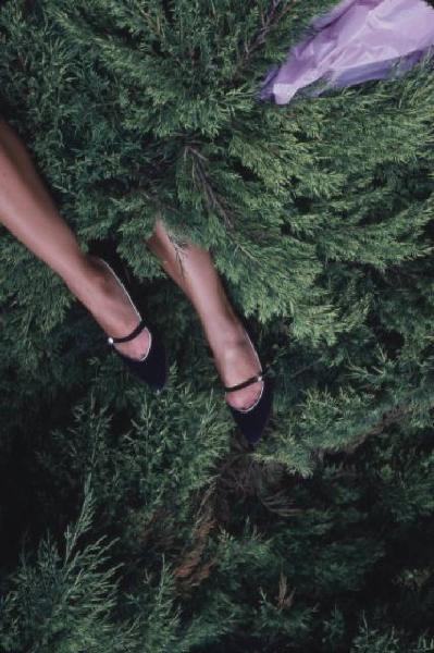 Gambe di donna con scarpa elegante nera sbucano da una fitta vegetazione - a margine un foulard lilla mosso dal vento