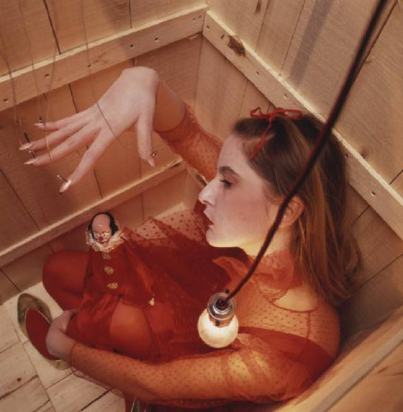 Ragazza in abito rosso chiusa in una scatola di legno. La ragazza tiene sulle braccia una bambola di porcellana