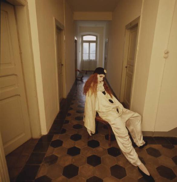 Ragazza in abito da Pierrot all'interno di una abitazione - la ragazza rimane appoggiata a una parete mentre da una delle porte che si affacciano sul corridoio fuoriesce una coppia di piedi