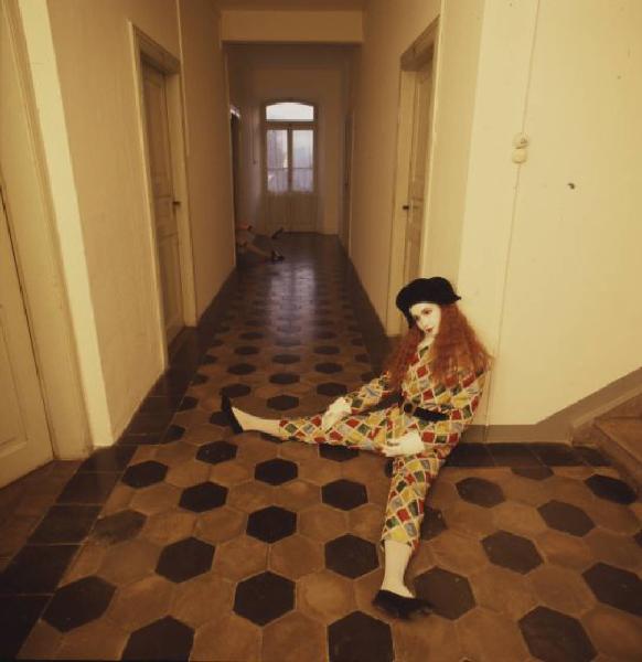 Ragazza in abito da Arlecchino all'interno di una abitazione - la ragazza rimane appoggiata a una parete mentre da una delle porte che si affacciano sul corridoio fuoriesce una coppia di piedi