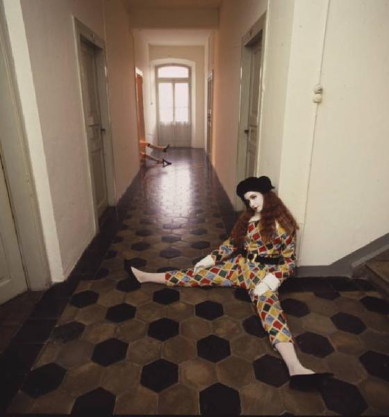 Ragazza in abito da Arlecchino all'interno di una abitazione - la ragazza rimane appoggiata a una parete mentre da una delle porte che si affacciano sul corridoio fuoriesce una coppia di piedi