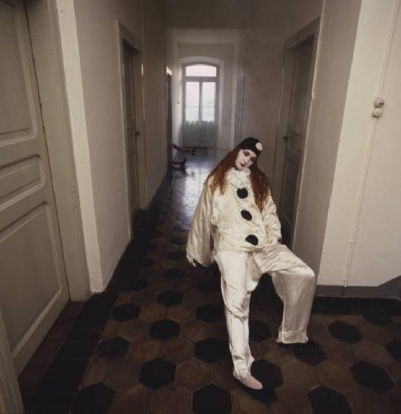 Ragazza in abito da Pierrot all'interno di una abitazione - la ragazza rimane appoggiata a una parete mentre da una delle porte che si affacciano sul corridoio fuoriesce una coppia di piedi