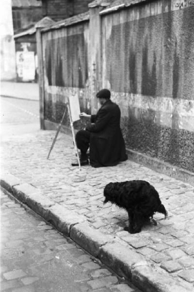 Parigi. Pittore al lavoro su un marciapiede di Montmartre. Accanto un cane