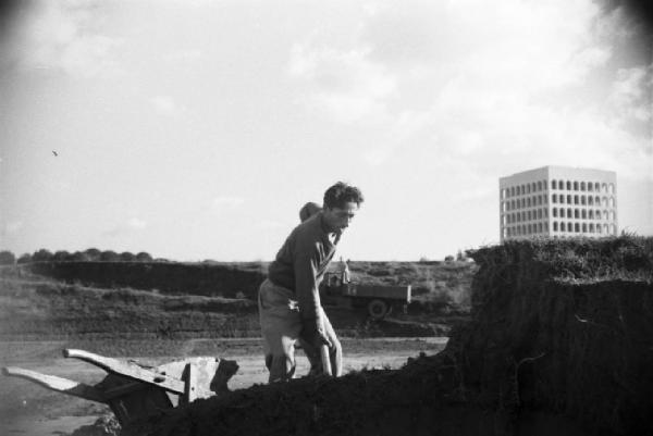 Italia Dopoguerra. Roma. Ponte sul Tevere in costruzione. Operaio dissoda il terreno munito di vanga e carriola. Sullo sfondo il Palazza della Civiltà e del Lavoro all'Eur