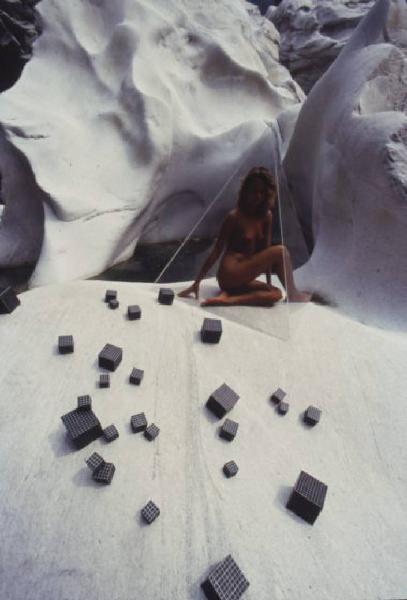 Nudo femminile - modella posa all'interno di una piramide in plexiglass. In primo piano alcuni cubi posizionati sulla roccia bianca e levigata