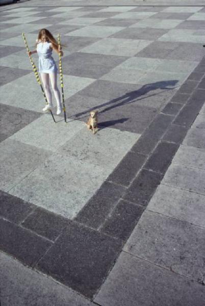 Modella sui trampoli in piazza con pavimentazione a scacchiera. Presenza di un cane di piccola taglia