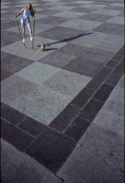 Modella sui trampoli in piazza con pavimentazione a scacchiera. Presenza di un cane di piccola taglia