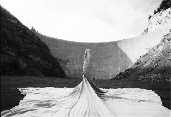 Figura umana avvolta in un lenzuolo sullo sfondo di una diga