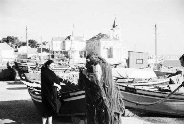 Cascais. Pescatore porta le reti in spalla sotto lo sguardo di una donna. In secondo piano imbarcazioni in secca