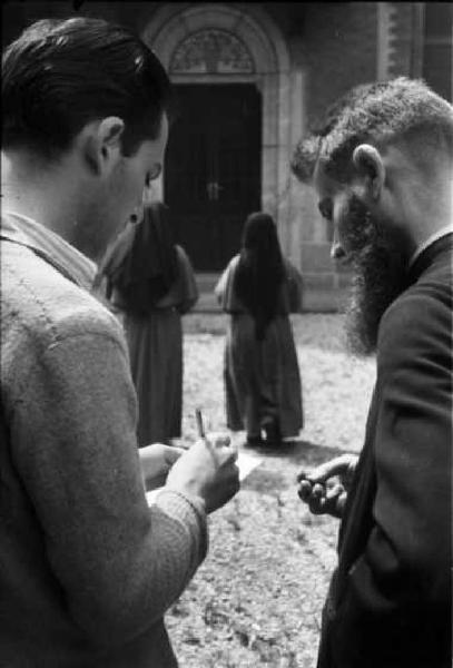 Un prete e un uomo con taccuino e matita di fronte all'ingresso di una chiesa - sullo sfondo due suore