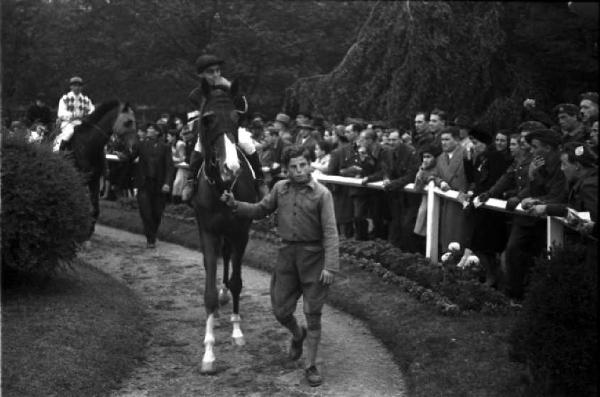 Italia Dopoguerra. Milano. Ippodromo di San Siro - Gran Premio. Folla di persone osserva fantini a cavallo mentre vengono accompagnati in pista