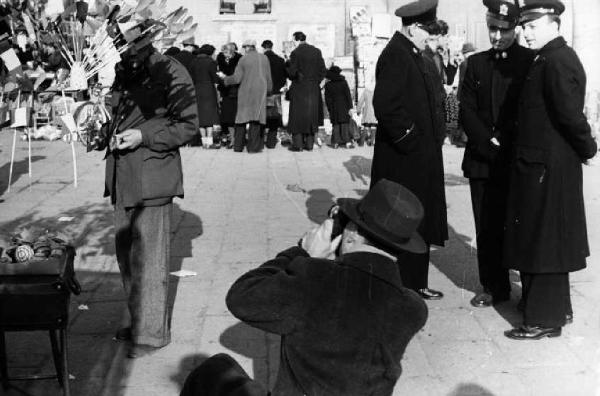 Italia Dopoguerra. Milano - fiera degli Obèi - Obèi. Fotografo riprende un ambulante; sulla destra un gruppo di poliziotti conversa