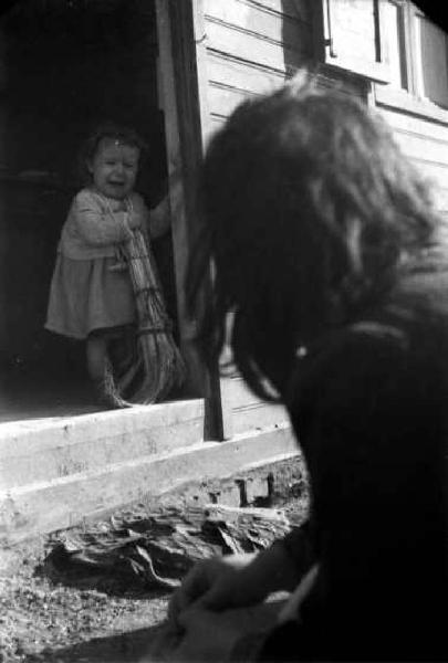 Italia Dopoguerra. Milano - Quartiere Baggio - Ritratto di bambino che piange sulla soglia di casa; la mamma di spalle lo osserva