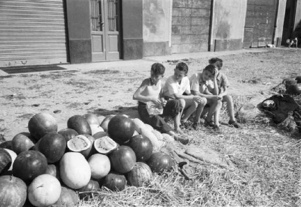 Italia Dopoguerra. Sant'Angelo Lodigiano. Reportage sulla figura di Santa Francesca Cabrini - gruppo di adolescenti seduti su un marciapiede chieccherano a fianco di un cumulo di angurie