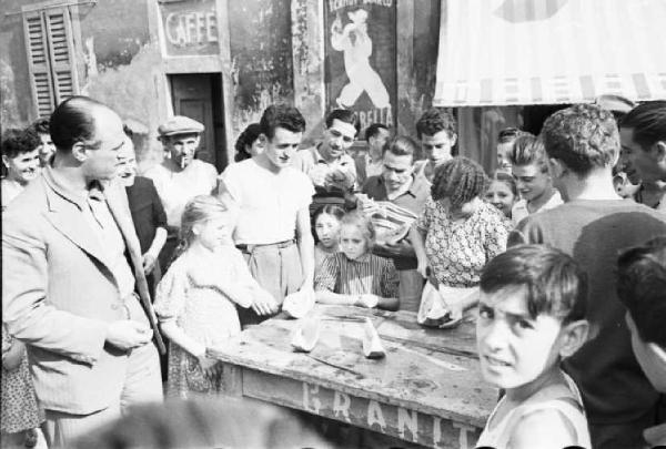 Italia Dopoguerra. Sant'Angelo Lodigiano. Reportage sulla figura di Santa Francesca Cabrini - un momento di vita del paese - in primo piano un gruppo di fanciulli attorno a un banchetto di legno per granite e angurie