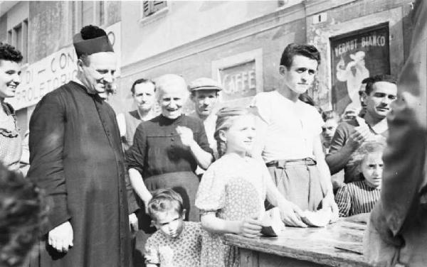 Italia Dopoguerra. Sant'Angelo Lodigiano. Reportage sulla figura di Santa Francesca Cabrini - un momento di vita del paese - in primo piano il parroco e un gruppo di fanciulli attorno a un banchetto di legno per granite e angurie