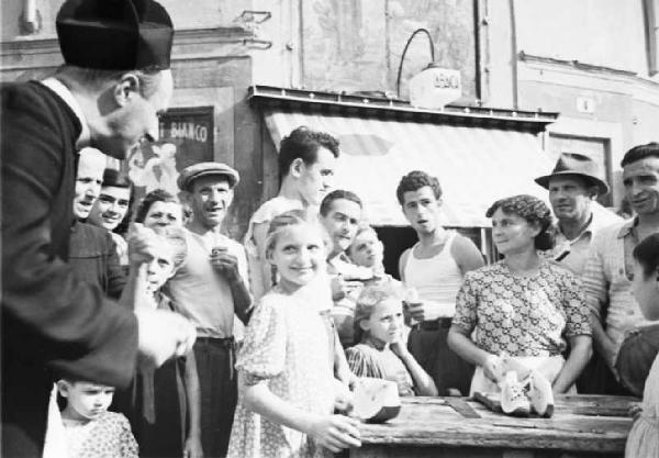 Italia Dopoguerra. Sant'Angelo Lodigiano. Reportage sulla figura di Santa Francesca Cabrini - un momento di vita del paese - in primo piano il parroco un gruppo di fanciulli attorno a un banchetto di legno per granite e angurie