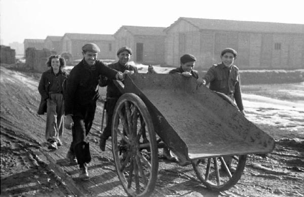 Italia Dopoguerra. Milano - Quartiere Baggio - Periferia - Edifici prefabbricati in legno - Terreno sterrato - Gruppo di ragazzi con carro