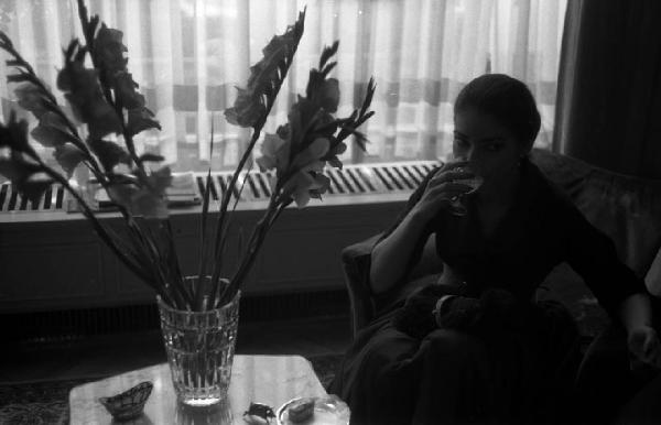 Milano - Abitazione di Maria Callas: interno - Ritratto femminile: Maria Callas (cantante lirica) seduta su una poltrona beve da un bicchiere - Tavolo con vaso di fiori