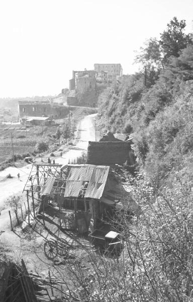 Italia Dopoguerra: Valmontone bombardata. Valmontone - Paesaggio: strada sterrata con baracche - Valmontone e Palazzo Doria in lontananza