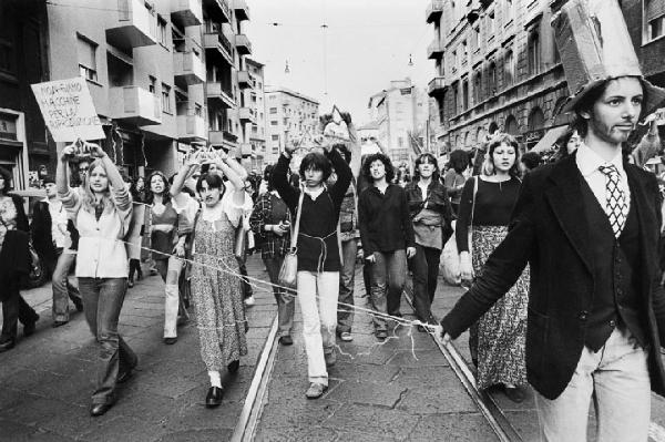 Azione teatrale alla manifestazione. Milano - Strada - Ritratto di gruppo: corteo di donne manifestanti, una travestita da uomo tiene legate con delle corde in vita altre tre donne - Cartello con scritta: "Non siamo macchine per la riproduzione"